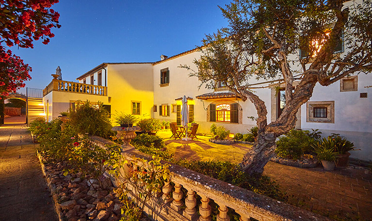 The Bennoc courtyard at dusk: a long sand-stone farmhouse with a balustraded terrace, warm light in the shuttered windows and an old olive tree in the foreground