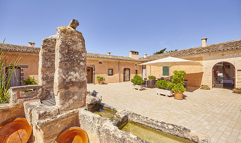 The Bennoc courtyard in full daylight: sand-coloured stone buildings on every side, a tiled terrace, potted plants and the low stone rim of an old fountain