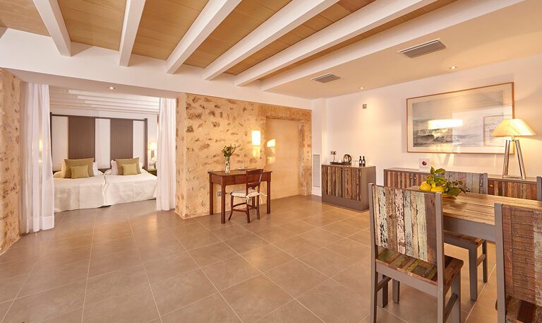A suite with a wall of bare natural stone behind a wide bed, a beamed and panelled ceiling, a writing desk and a small dining table of reclaimed wood