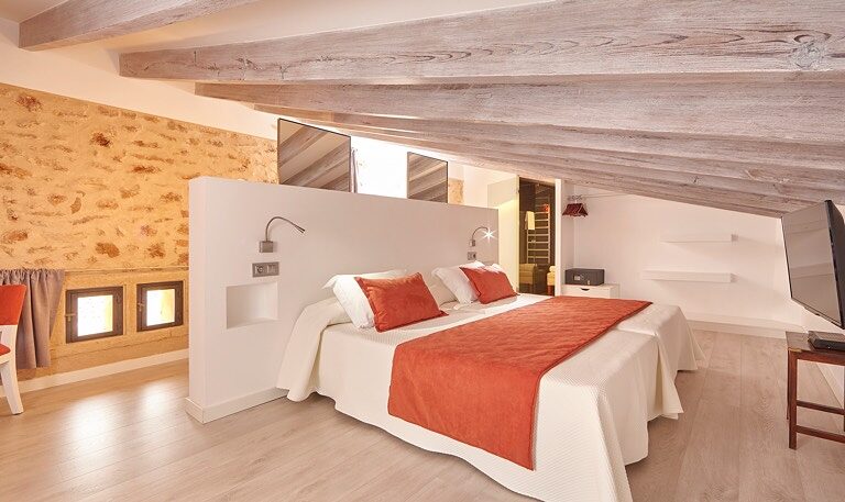 A double room under a whitewashed log-beam ceiling, an exposed stone wall behind the bed, white linen with a terracotta runner and two small shuttered windows